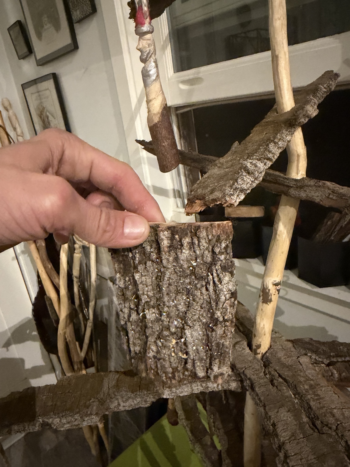 A hand holds a rectangular bark fragment in front of an unfinished tree form with bark tiers and a wood-round base.